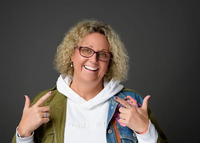 Anna Bates smiling and pointing towards herself, wearing glasses and a green jacket.